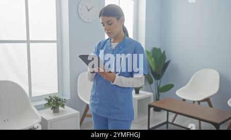 Focused young woman nurse using a tablet in a bright hospital room with modern furniture and plants Stock Photo
