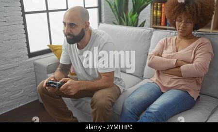 An interracial couple in a living room, man playing video games and woman upset, depicting relationship issues or leisure time conflicts. Stock Photo