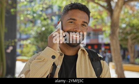A smiling young black man with a beard engaging in a phone conversation outdoors in an urban park setting. Stock Photo