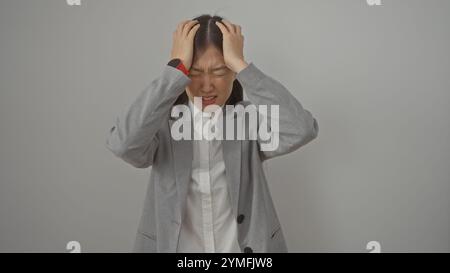 An asian woman in a gray suit with hands on her head against a white background, showing stress or headache Stock Photo
