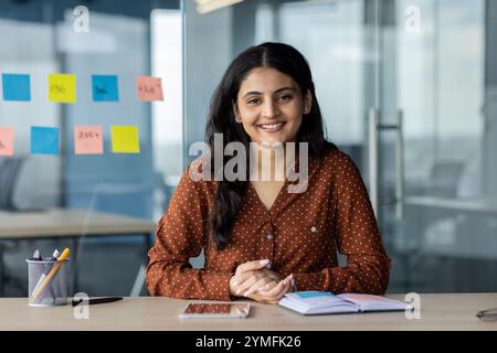 Latin American businesswoman smiling at desk in modern office. Confident, professional ambiance with smartphone, notebook. Represents success, motivation, casual work environment. Stock Photo