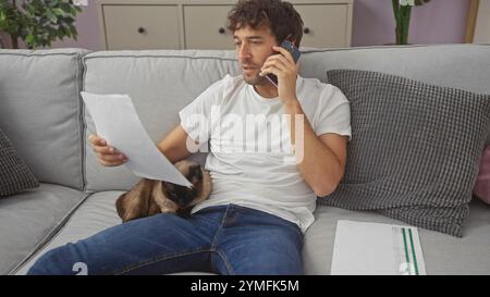 A hispanic man engages in a phone conversation while examining documents and enjoying the company of a siamese cat on a couch indoors. Stock Photo