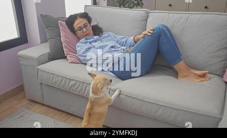A middle-aged woman relaxes on a couch in a cozy living room, interacting with her attentive chihuahua pet Stock Photo