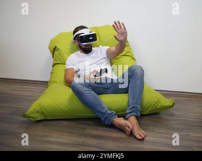 Man is seated on green bean bag, using VR headset and holding controller Stock Photo