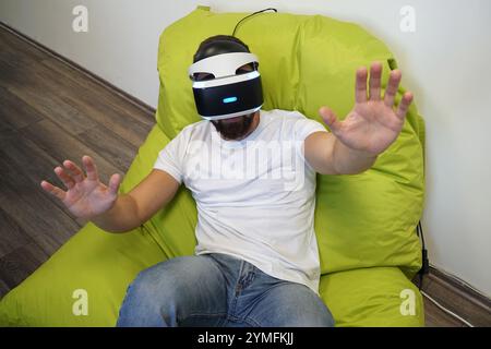 Man seated on green bean bag, wearing VR headset, extending his hands in virtual environment Stock Photo
