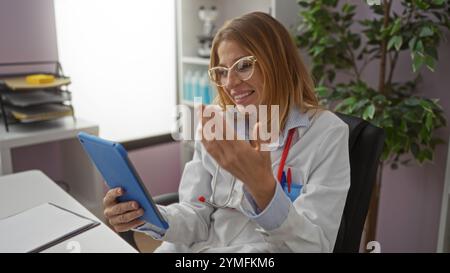 Woman doctor video calling in a hospital office, holding a tablet, smiling and engaging with a colleague or patient, highlighting modern healthcare co Stock Photo