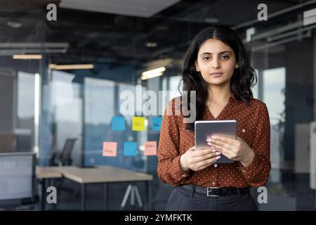 Latin American woman businesswoman with tablet in modern office setting. Confident, focused professional showcasing technology in workplace environment with glass walls and sticky notes. Stock Photo