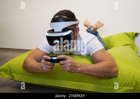 Man lying face down on green bean bag, fully immersed in VR gaming session with controller Stock Photo