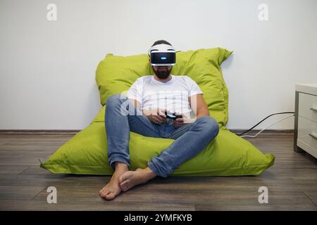 Man wearing VR headset, sitting comfortably on green bean bag while gaming with controller Stock Photo