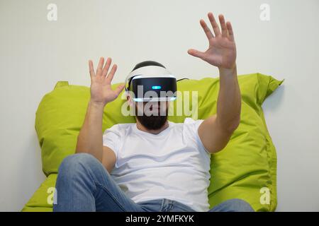 Man seated on green bean bag, wearing VR headset, extending his hands in virtual environment Stock Photo