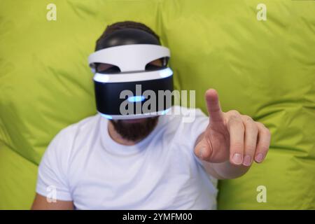 Man seated on green bean bag, wearing VR headset, extending his hands in virtual environment Stock Photo