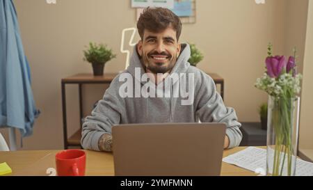 Handsome young hispanic man with a beard smiling while working on a laptop in a cozy home interior. Stock Photo