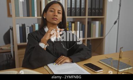 Young hispanic female judge wearing a black robe in an office setting, holding glasses with a confident expression, surrounded by paperwork, books, an Stock Photo