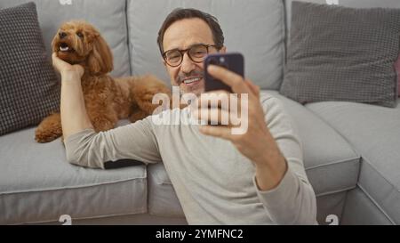 A smiling middle-aged man takes a selfie with a poodle in a cozy living room setting. Stock Photo