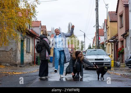 Three cheerful women interacting with curious black cat on quaint cobblestone street with colorful houses and autumn leaves. Concept of friendship, ur Stock Photo