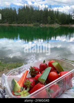 A scenic view of a clear lake surrounded by trees, with a close-up of fresh strawberries, cucumbers, and cherry tomatoes in a plastic container. Ideal Stock Photo