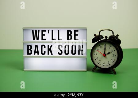 We'll Be Back Soon text on LED lightbox with alarm clock on green background Stock Photo