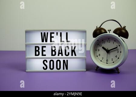 We'll Be Back Soon text on LED lightbox with alarm clock on purple background Stock Photo