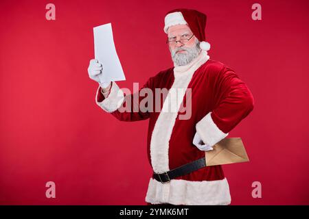 Portrait of Santa Claus standing against red background, holding paper in one hand and envelope in other, wearing traditional red and white outfit Stock Photo