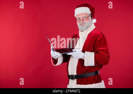 Santa Claus wearing red suit and hat standing against red background, holding and using laptop while smiling. Santa Claus showing festive enjoyment and connecting with technology Stock Photo