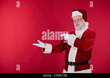 Smiling Santa Claus in red suit with white beard pointing with cheerful expression in front of red background. Ample copy space on red background for holiday text or designs Stock Photo