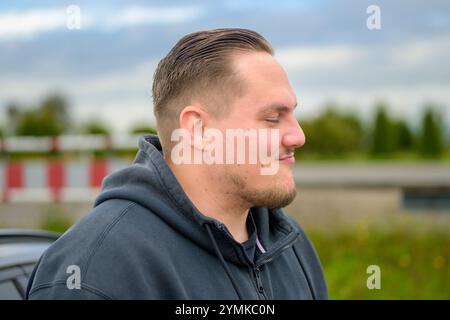 A man with a neatly groomed short haircut and facial stubble stands in profile, eyes closed, wearing a dark hoodie. Stock Photo