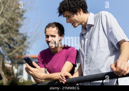 Two men, a gay couple, are using a smartphone and smiling while leaning on a railing outdoors Stock Photo