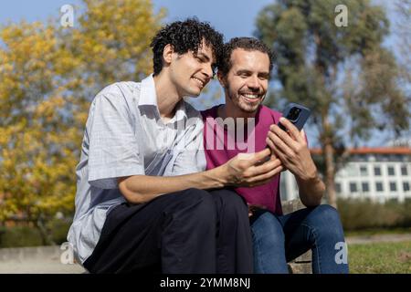 Two men are sitting outdoors, smiling, and using a mobile phone together Stock Photo