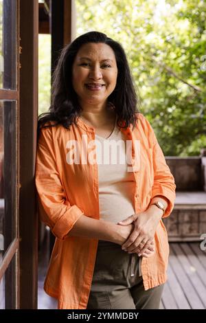 Smiling senior woman in orange shirt standing on porch, enjoying sunny day. Outdoors, sunshine, happiness, summer, relaxation, cheerful Stock Photo