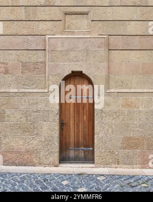 A weathered wooden door stands closed against a textured stone wall, framed by soft hues of beige and gray on a Cobblestone Street in a Historic Area Stock Photo