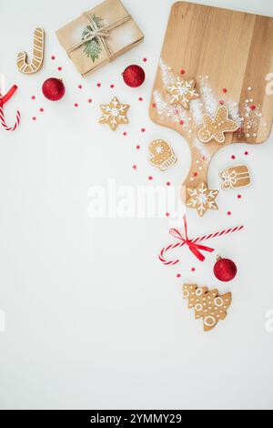 Top view of Christmas gingerbread cookies on a wooden board, red baubles, candy canes, a wrapped gift, and festive decorations scattered on a white ba Stock Photo