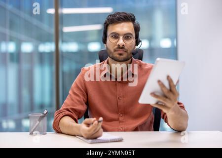 Latin American man businessman in office using digital tablet and headset for video call. Engaged in remote communication, showcasing modern corporate technology and connectivity. Stock Photo