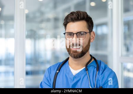 Young man doctor in blue scrubs and glasses in hospital corridor, exuding confidence and professionalism. Ideal for showcasing healthcare professional themes. Smiling with stethoscope. Stock Photo