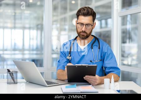 Man doctor wearing blue scrubs and glasses reviewing documents with laptop and stethoscope in modern office. Professional healthcare setting with focused attention on medical paperwork. Stock Photo