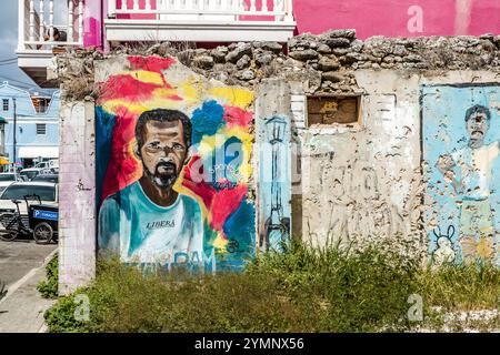 Portrait mural on a heavily weathered rubble-stone wall in Breedestraat, Willemstad, Curaçao. The mural depicts a person wearing a T-shirt bearing the word ‘LIBERA’ against a colourful, expressive background; cracks, flaking plaster and exposed masonry are deliberately incorporated into the work by the street artists as design elements. Cracks, peeling plaster and exposed brickwork are not covered up, but incorporated into the artworks as design elements. Kòrsou Stock Photo
