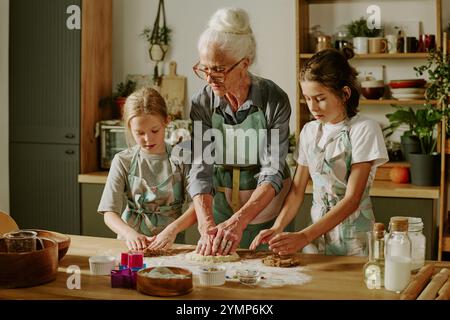 Senior woman baking with two children in kitchen, wearing matching aprons and making cookies on wooden countertop Stock Photo