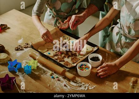 Children decorating holiday cookies in modern kitchen with various cookie cutters and colorful decorations placed on wooden table, creating festive atmosphere Stock Photo