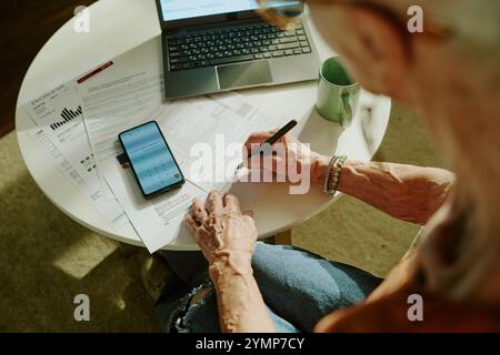 Senior woman seated at table, using smartphone and laptop while writing on financial documents. Engaging with technology as part of daily tasks Stock Photo