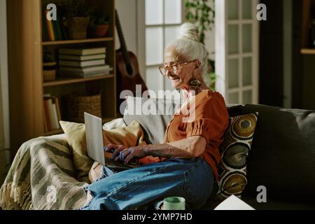 Senior woman sitting on sofa looking at laptop screen in cozy living room filled with books and cushions while wearing glasses and colorful clothes Stock Photo