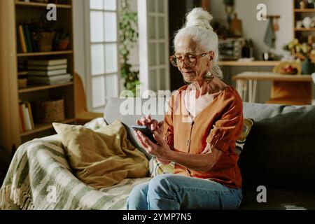 Elderly woman sitting on couch in cozy living room, using smartphone for browsing internet, with shelves and kitchen in background Stock Photo