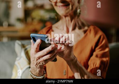 Close-up of elderly woman with light grey hair and glasses using smartphone while smiling. Senior is wearing casual outfit and sitting on couch in well-lit living room Stock Photo