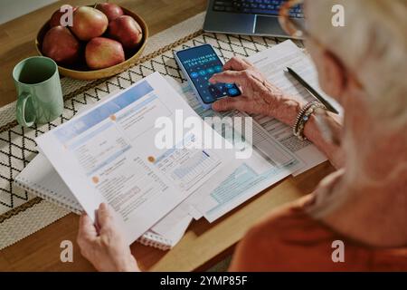 Close-up of elderly person sitting at table, examining financial documents while using smartphone calculator. Visible laptop, green mug, and apples enhance the work environment Stock Photo