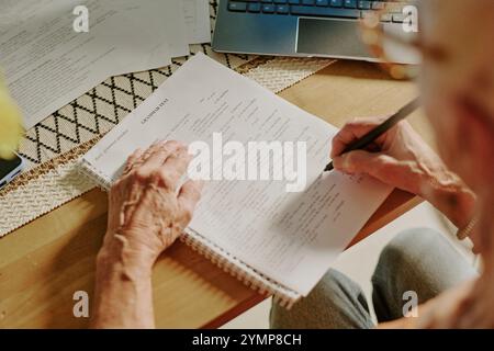 Senior person carefully writing on paper, with laptop open on table, creating organized workspace, focusing on handwriting, surrounded by documents Stock Photo