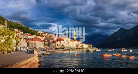 The view оf Perast, Montenegro, illuminated by the warm, golden light of the setting sun. The foreground is serene, with boats floating, while dark st Stock Photo