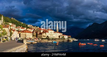 The view оf Perast, Montenegro, illuminated by the warm, golden light of the setting sun. The foreground is serene, with boats floating, while dark st Stock Photo