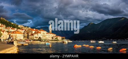 The view оf Perast, Montenegro, illuminated by the warm, golden light of the setting sun. The foreground is serene, with boats floating, while dark st Stock Photo