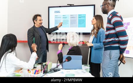 Multigenerational diverse business team in meeting room discussing content planning strategy while senior manager presents weekly timeline on large sc Stock Photo