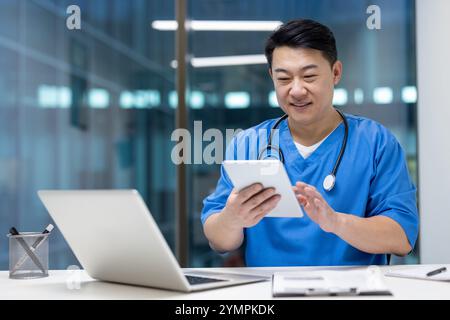 Asian man doctor using tablet and laptop in office, showcasing efficiency and modern healthcare solutions. Wearing scrubs, smiling, focused on digital screen, enhancing medical productivity. Stock Photo