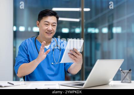 Asian man doctor in blue scrubs using tablet for virtual consultation in office. Engaged in video call, smiling confidently, with laptop on desk, conveying professionalism and technology. Stock Photo