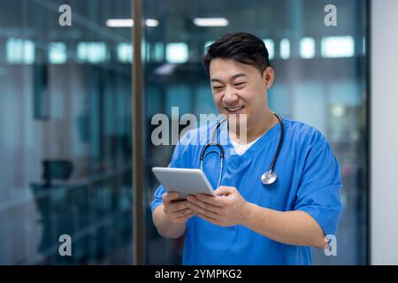 Asian man doctor in scrubs using tablet computer, smiling while checking medical records. Technology in healthcare enhances patient care, facilitating communication within medical team. Stock Photo